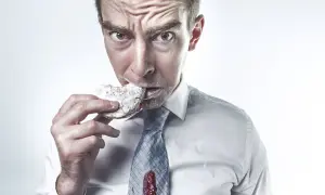 Mężczyzna w białej koszuli i krawacie, z plamą dżemu, je pączka. Jego twarz wyraża frustrację, jakby walczył z głodem i hormonem grelina.