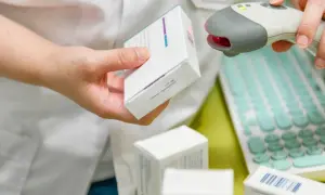 Farmaceuta skanuje lek, pokazując, ile ważna jest recepta. W tle klawiatura i inne opakowania.