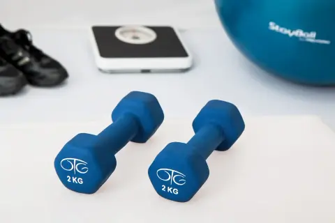 Dwa niebieskie hantle po 2 kg, waga, buty sportowe i piłka gimnastyczna.