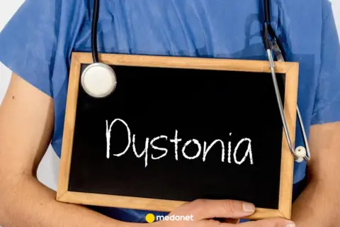 Lekarz w niebieskiej koszuli trzyma tabliczkę z napisem "Dystonia". Stetoskop zwisa na jego ramieniu.