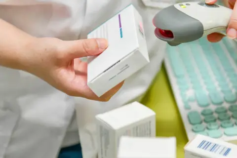 Farmaceuta skanuje lek, pokazując, ile ważna jest recepta. W tle klawiatura i inne opakowania.