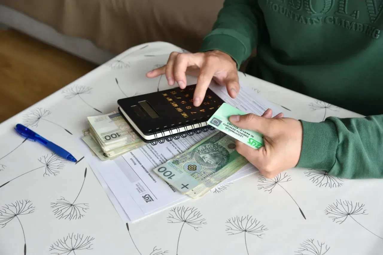 Ręce liczą pieniądze i dokumenty ZUS. Na stole leżą polskie banknoty, kalkulator i karta legitymacyjna emeryta-rencisty, co sugeruje sprawy związane z rentą chorobową.