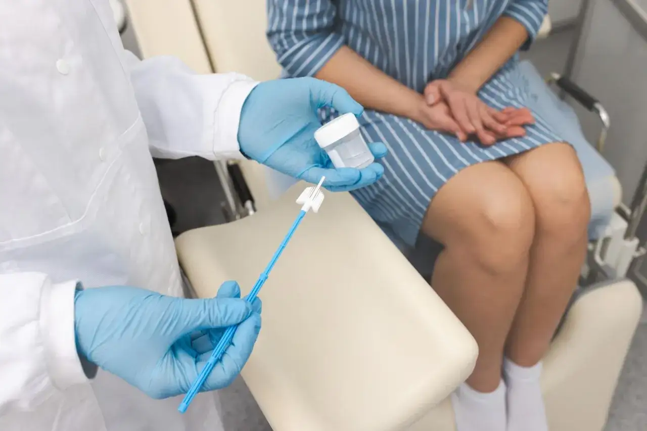 Lekarz w niebieskich rękawiczkach trzyma pojemnik i szczoteczkę do pobrania wymazu. Pacjentka siedzi w fotelu ginekologicznym, przygotowując się do badania. To ważny krok w profilaktyce zdrowia, zwłaszcza w kontekście badań cytologicznych, które pomaga...